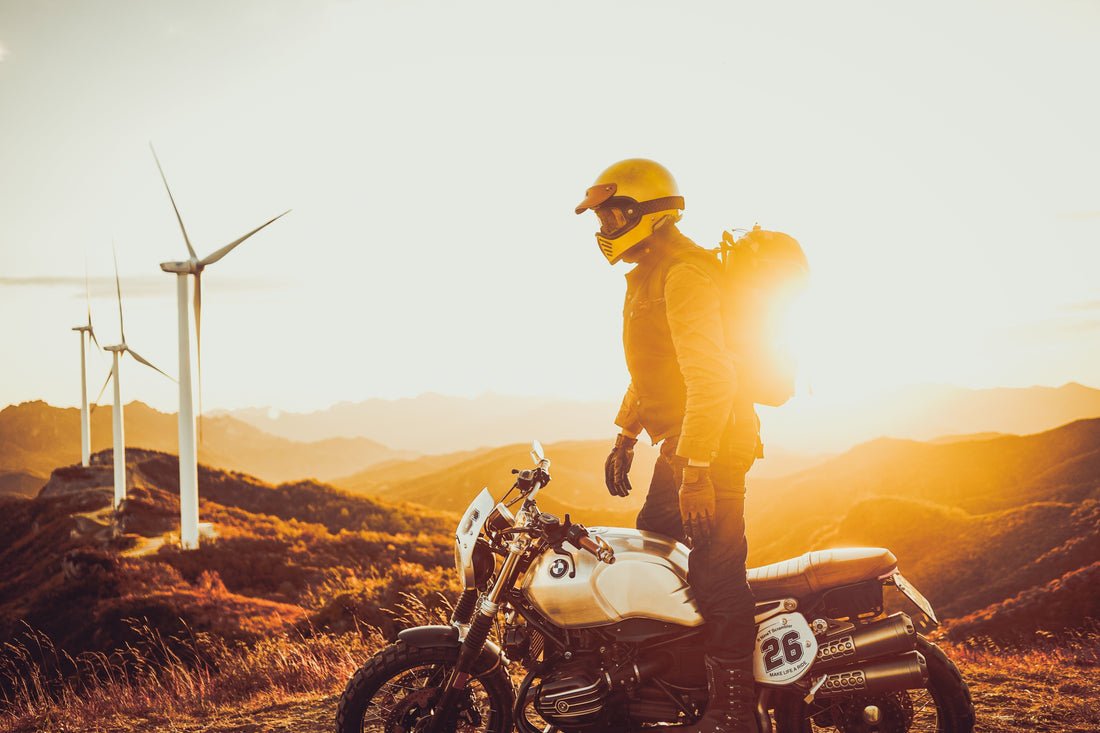 Rider with yellow helmet and backpack standing by a motorcycle at sunset blog cover