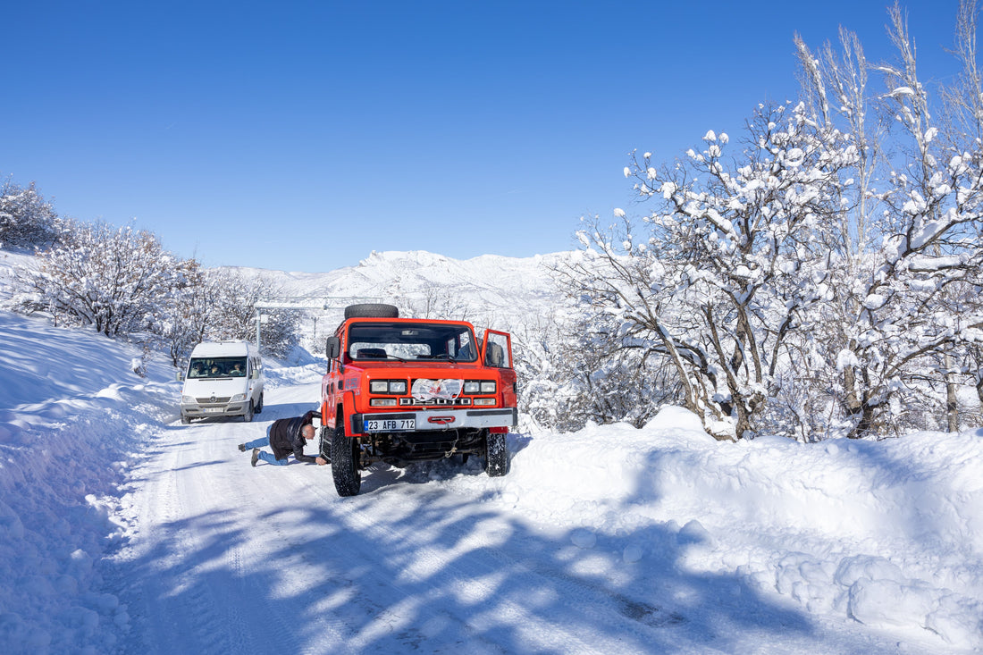 Driver lying on snowy road repairing underside of red off-road vehicle in winter blog cover